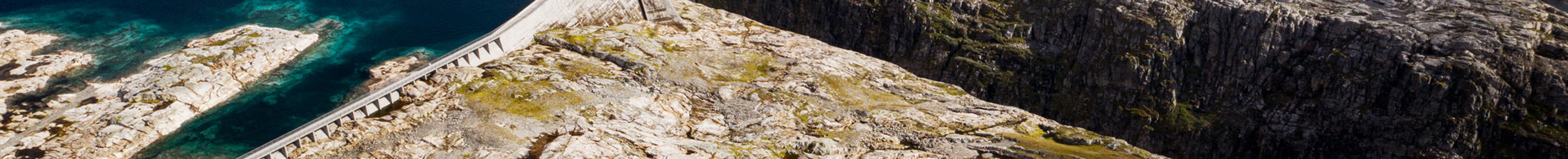 Diga di cemento curvo che attraversa un paesaggio roccioso, creando un lago blu, rocce grigie con vegetazione sparsa.